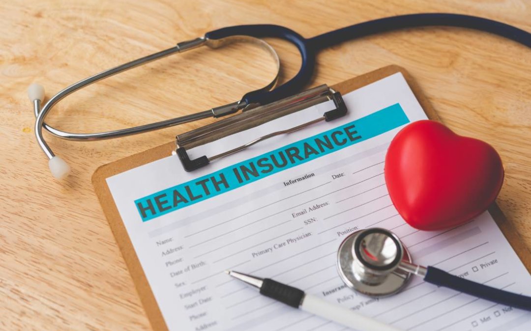 Health Insurance Form on a clipboard, near pen, stethoscope, and foam heart.