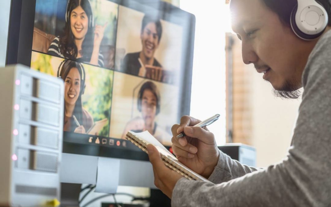 A remote worker takes notes while on his video call with four other colleagues.